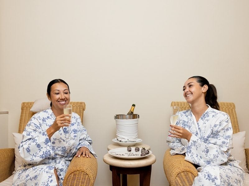 Two women in white robes sitting in wicker chairs holding glasses of champagne